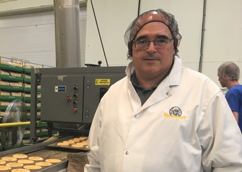 A man wearing a long white coat and a hairnet standing next to a piece of industrial equipment with a cookie sheet on it.