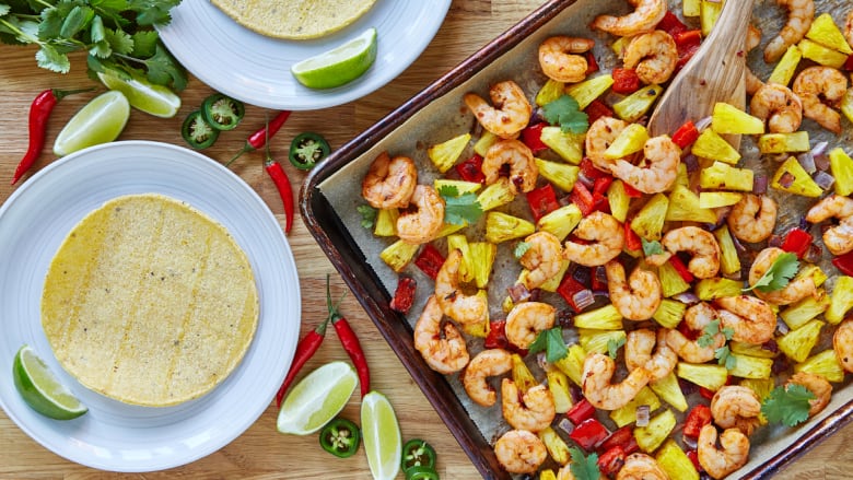 Overhead shot of a sheet pan on a wooden table. The pan has roasted shrimp and pieces of pineapple on it. 2 white plates with tortillas on them sit next to the pan. 