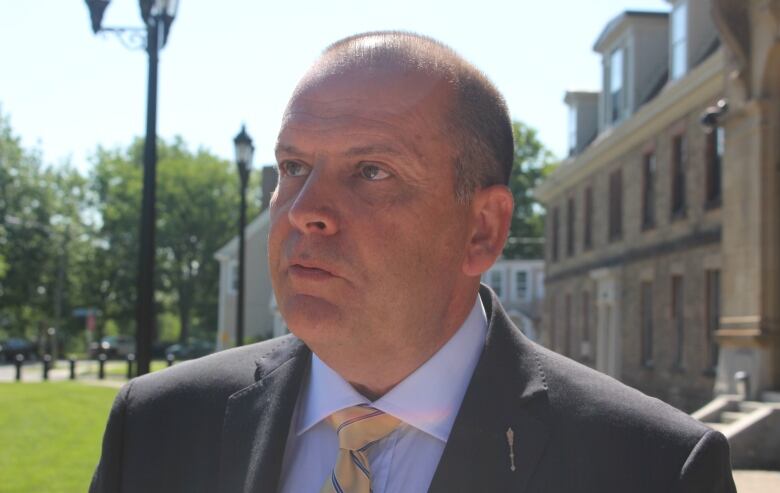 Balding man in suit and tie standing outside on a sunny day.