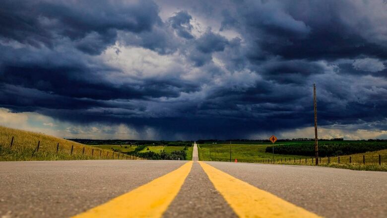 Two yellow lines run through the centre of a rural highway. Dark clouds can be seen ahead.