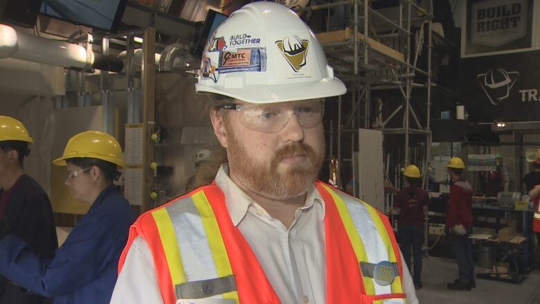 A white man with red hair is seen wearing a hard hat, safety goggles and a reflective vest.