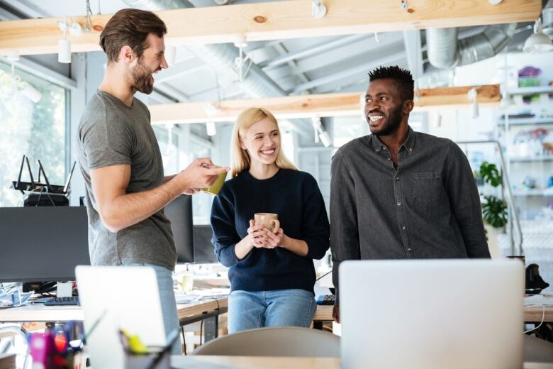 Three people laugh and smile together while holding mugs and standing in an office. 