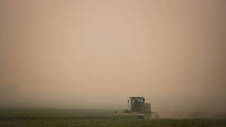 A tractor is running through a field. The sky is very cloudy and tinged orange from wildfire smoke. 