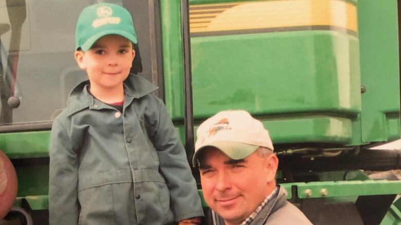 A young boy in coveralls stands on a farm vehicle, standing alongside a man kneeling.