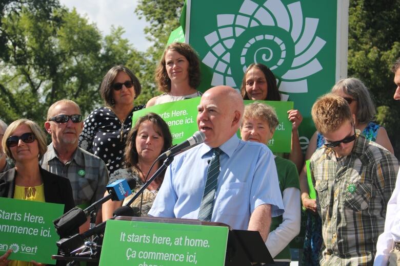 A man speaking into a microphone in front of a group of people holding green signs.