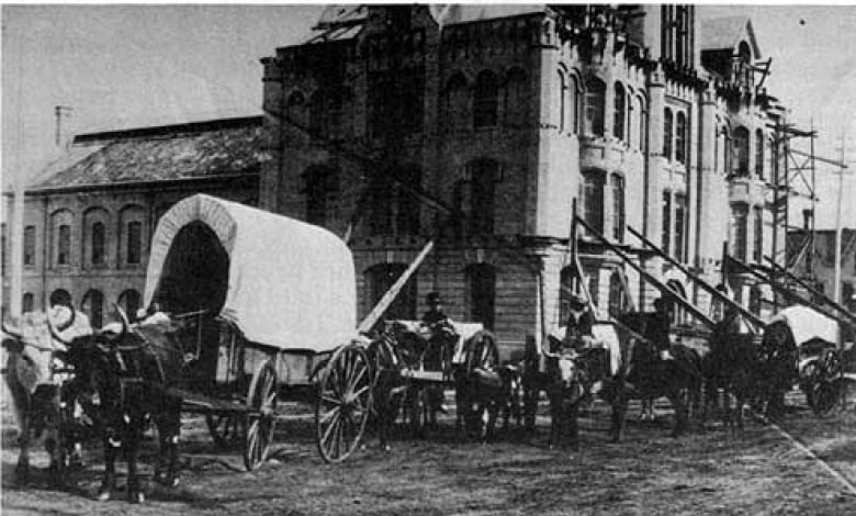 Horses and carts and people stand around an old building on a mud street.