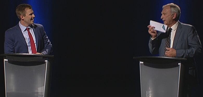 Two men standing at podiums face each other, speaking.