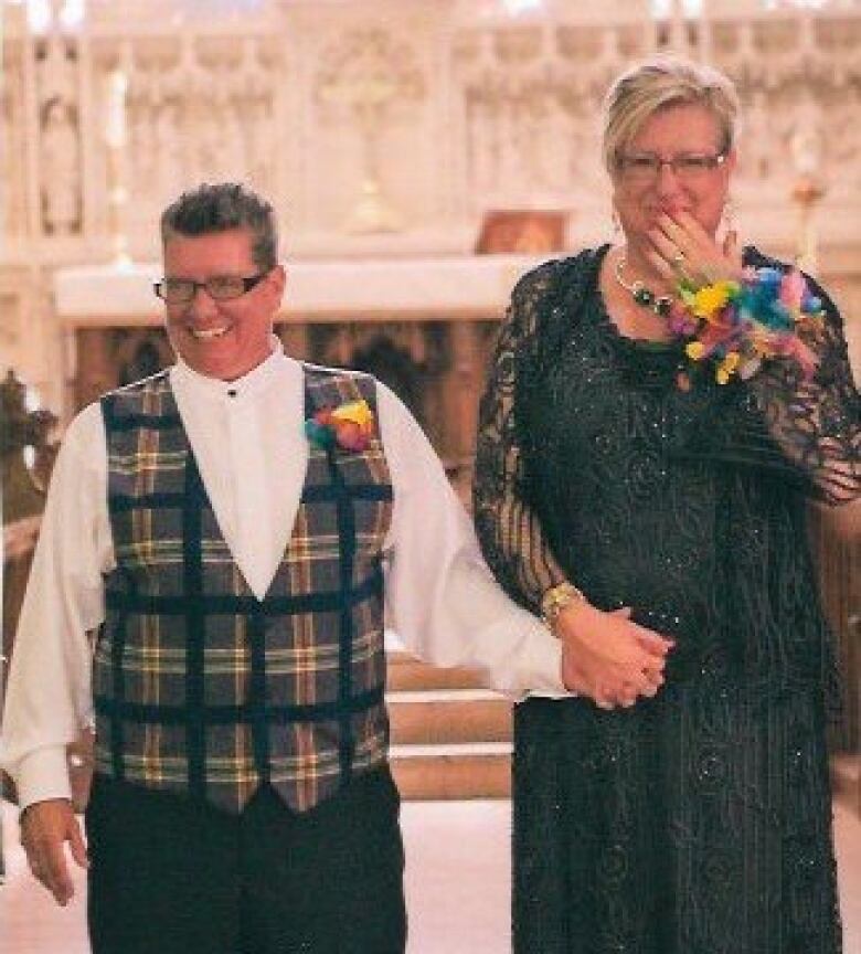 Two women, one in a dress and the other in a dress shirt and vest, smile at the front of a church. They are both wearing corsages.