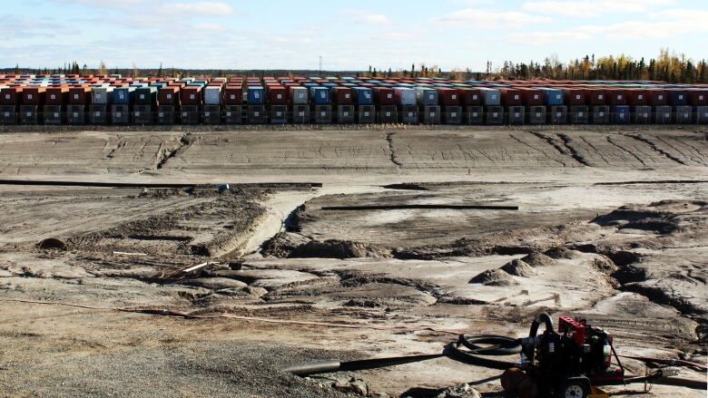 Rows of shipping containers sit on top of each other in the distance on a landscape that is sand and dirt. There is some machine that sits in the front.