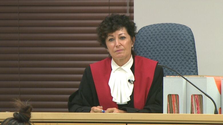 A woman in a black robe with a red band sits behind a judge's bench.