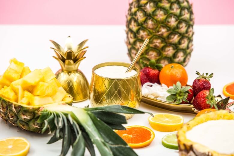 A golden pineapple-shaped cup with a white dessert in it on a pink surface with pink background. A hollowed-out pineapple with pineapple chunks in it, orange, lemon and lime slices, a full pineapple, and a tray of strawberries sit around the cup. 