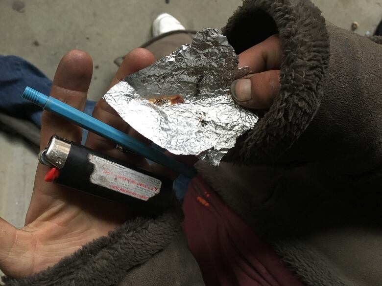 A person holds up a piece of aluminum foil with a brown substance on it, along with a lighter and a small pipe.