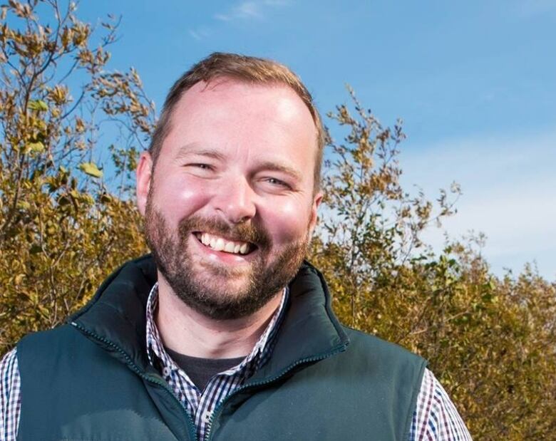 A smiling man stands before trees and green space.