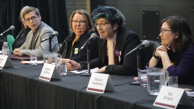 Five people sit at a table with microphones in front of them. One of them is leaning in to speak. 