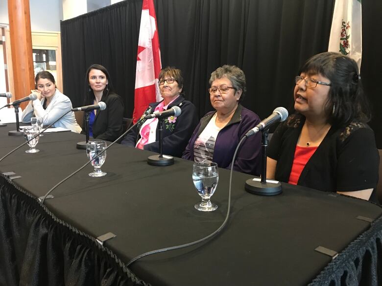 Five women sit in a row behind a table, with microphones set up in front of them.