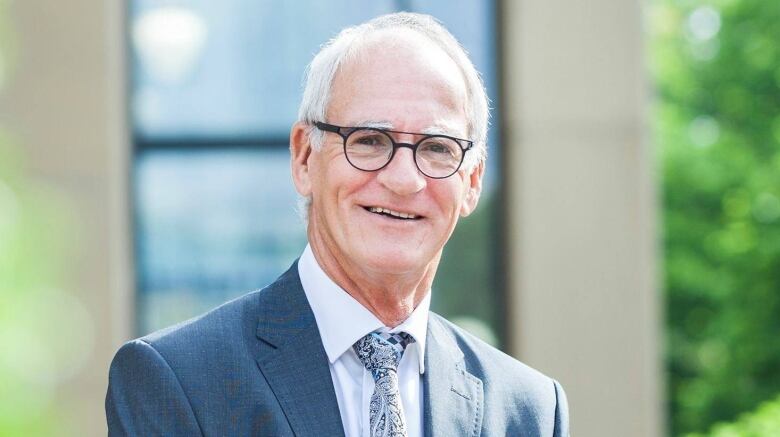 A white haired man with glasses wear a suit and stands outdoors, smiling at the camera.