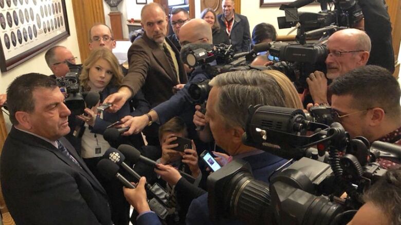 A group of reporters with microphones and cameras surround a man in a suit. 