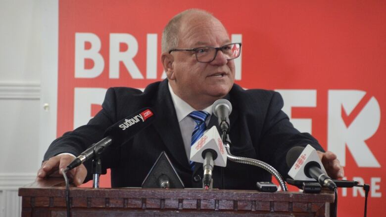 A balding man with glasses wearing a suit speaks into microphones at a podium.