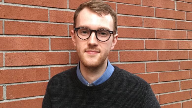 A man with short hair and dark rimmed glasses standing in front of a red brick wall