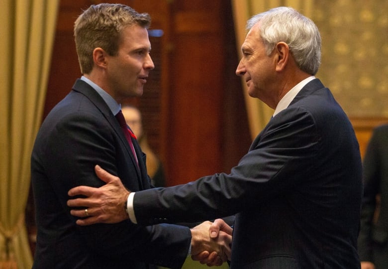 Two men shake hands. The man on the left puts his left hand on the man's upper arm while shaking with his right.