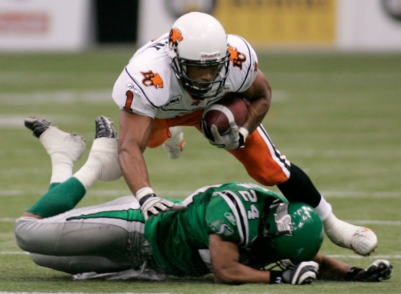 Josh Boden carries a football and wears a white B.C. Lions jersey as he trips over a green-clad Saskatchewan Roughriders player.