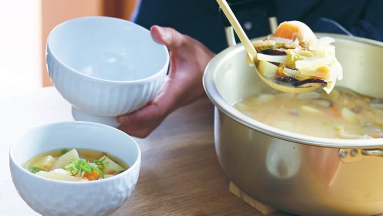 Closeup on someone ladling miso soup with root vegetables from a silver pot into two white bowls. 