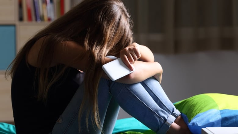 A girl sits with her knees drawn up under her chin and her hands, which hold a cellphone, hiding her face.