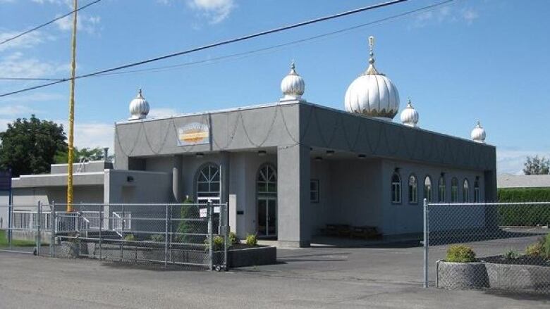 A grey one story gurdwara building with five visible white and golden spires consistent with Sikh places of worship.