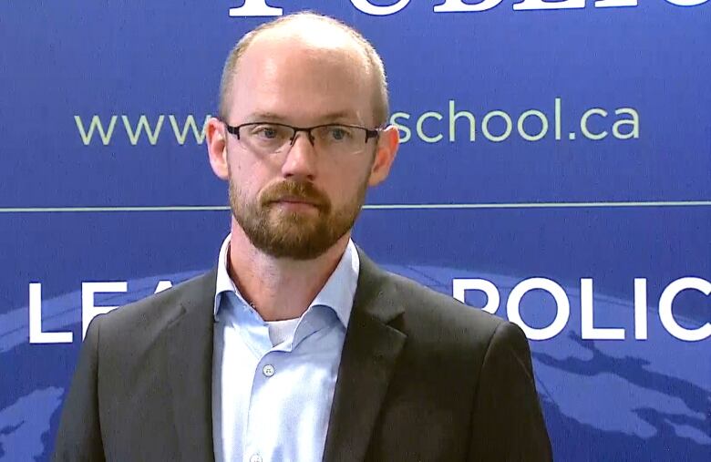 Trevor Tombe is seen wearing a suit jacket and a blue-collared shirt. He's standing in front of a blue backdrop.