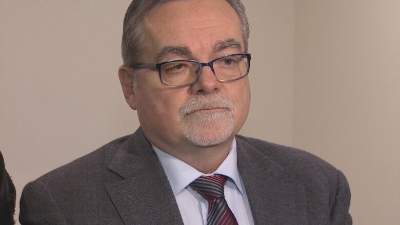 A man with glasses wearing a grey suit and grey and red striped tie.