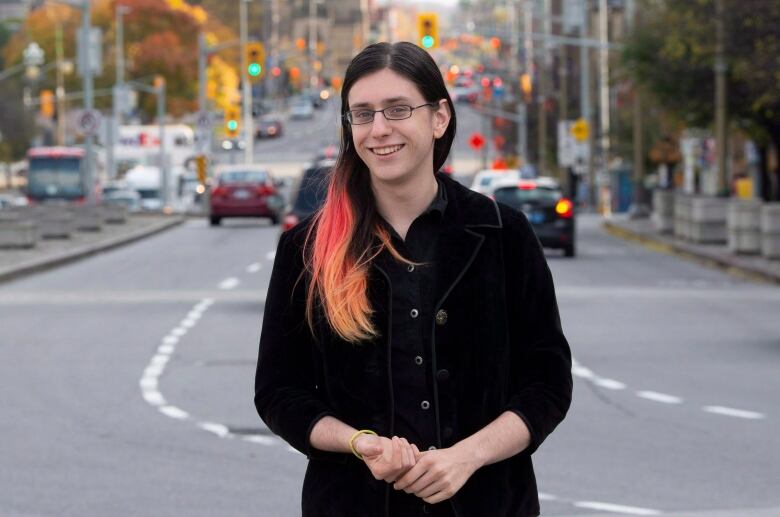 A woman in black with glasses and pink highlights in her hair smiles with an urban streetscape in the background.