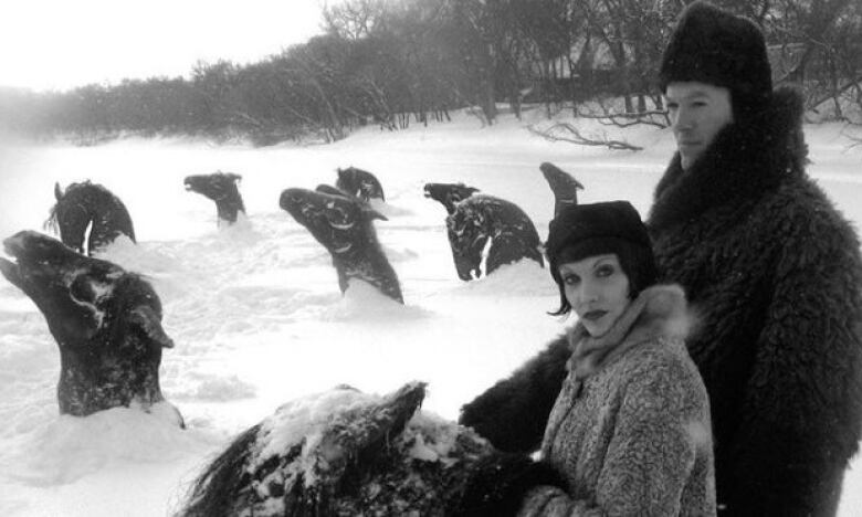 A black and white still from a film shows two people in fur coats standing next to a frozen lake, with the heads of frozen horses protruding from the ice.