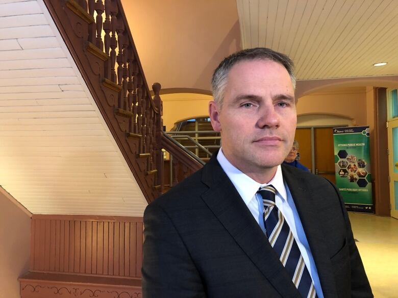 A middle-age man in a suit and tie stands near the interior stairs of an older-looking building.