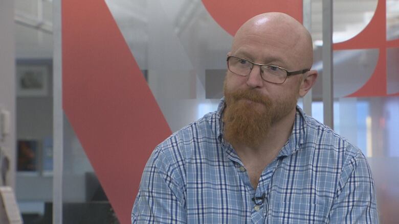 A bald man with a red beard in a blue plaid collared shirt sits for an interview with a CBC television studio set behind him.