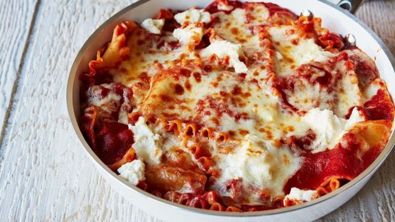 A skillet on a grey wood surface. The skillet is filled with a cooked lasagna with melted cheese and dollops of ricotta cheese on top. 