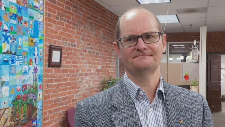 A man in a suit jacket and button-up shirt stands in front of an interior brick wall in an office. 