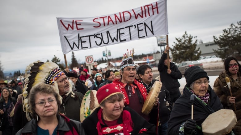 A group of people, some in First Nation's regalia and carrying drums, march beneath a sign saying 'We Stand with Wet'suwet'en.'