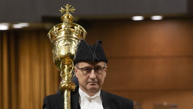 A man wearing a black suit and a traditional House of Commons white collar carries a gold ceremonial mace into the House.