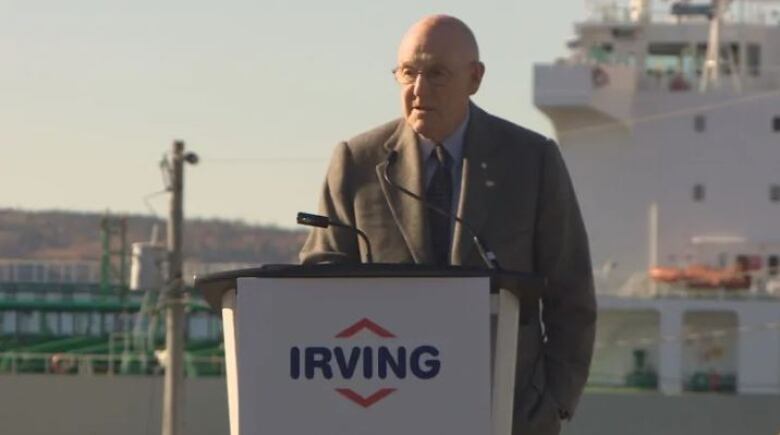 An older man speaking at a stand outdoors. The stand has a microphone on it and the front has a logo that says 