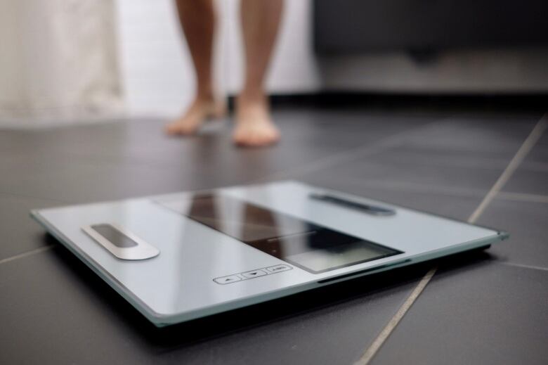 A white electronic bathroom scale sits on a floor with a woman's legs in the background.