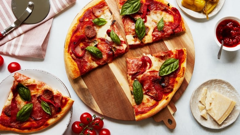 Overhead shot of a circular pizza with meat, cheese and fresh basil on top. It's sitting on a wooden cutting board and one of its slices is next to it on a white plate. A pizza cutter and a bowl of Parmesan cheese are next to the pizza. 