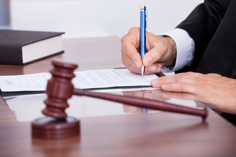 A close-up photo shows a gavel on a desk and the hands of a man writing in a document.