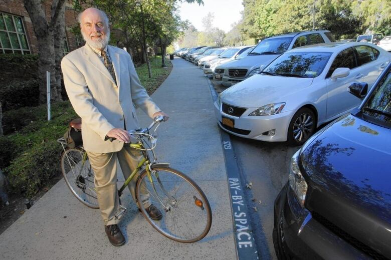 An older man in a jacket and tie with his bike beside a row of parked cars.