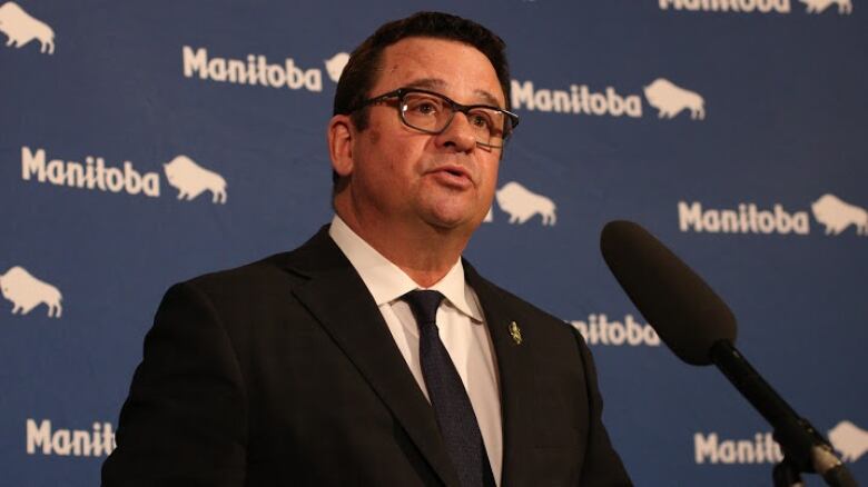 A man with glasses in a suit and tie speaks into a microphone in front of a Manitoba government backdrop.