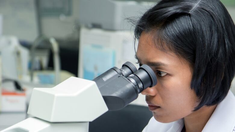 A woman wearing a white lab coat and white latex gloves peers into a large microscope in a laboratory.