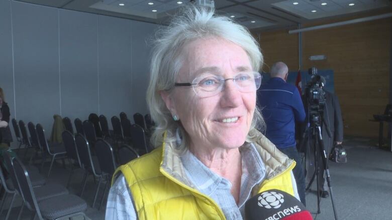 A woman speaking into a CBC microphone. She is wearing a yellow vest.