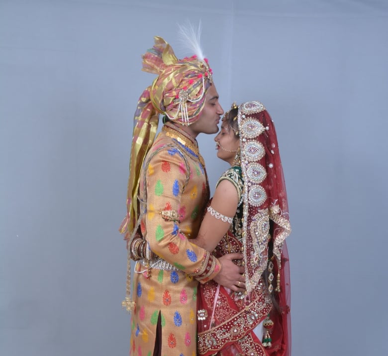 A groom kisses the bride's forehead as they embrace. They are wearing traditional Indian wedding attire.  