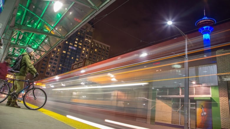 A long-exposure photo of an LRT train passing a station platform, with a man on a bicycle watching it pass.