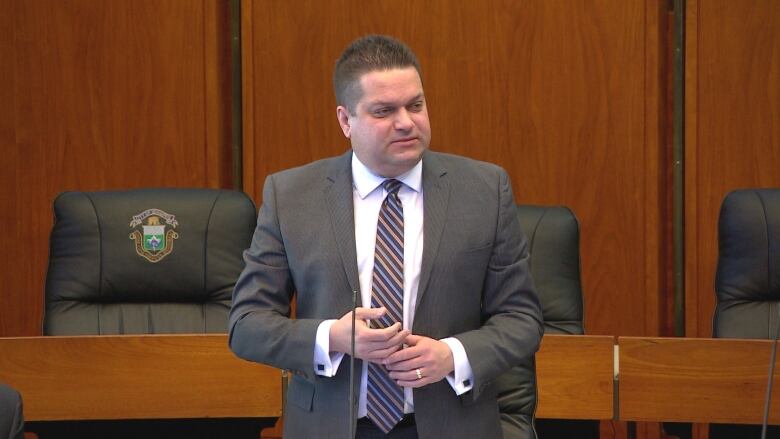 A man wearing a suit is standing in a room with chairs and desks behind him.