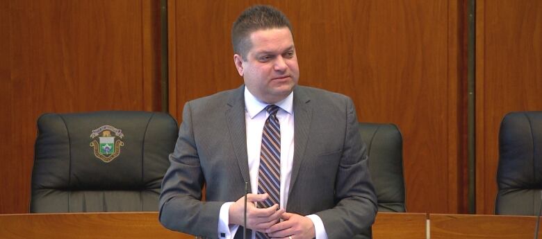 A man wearing a suit is standing in a room with chairs and desks behind him.